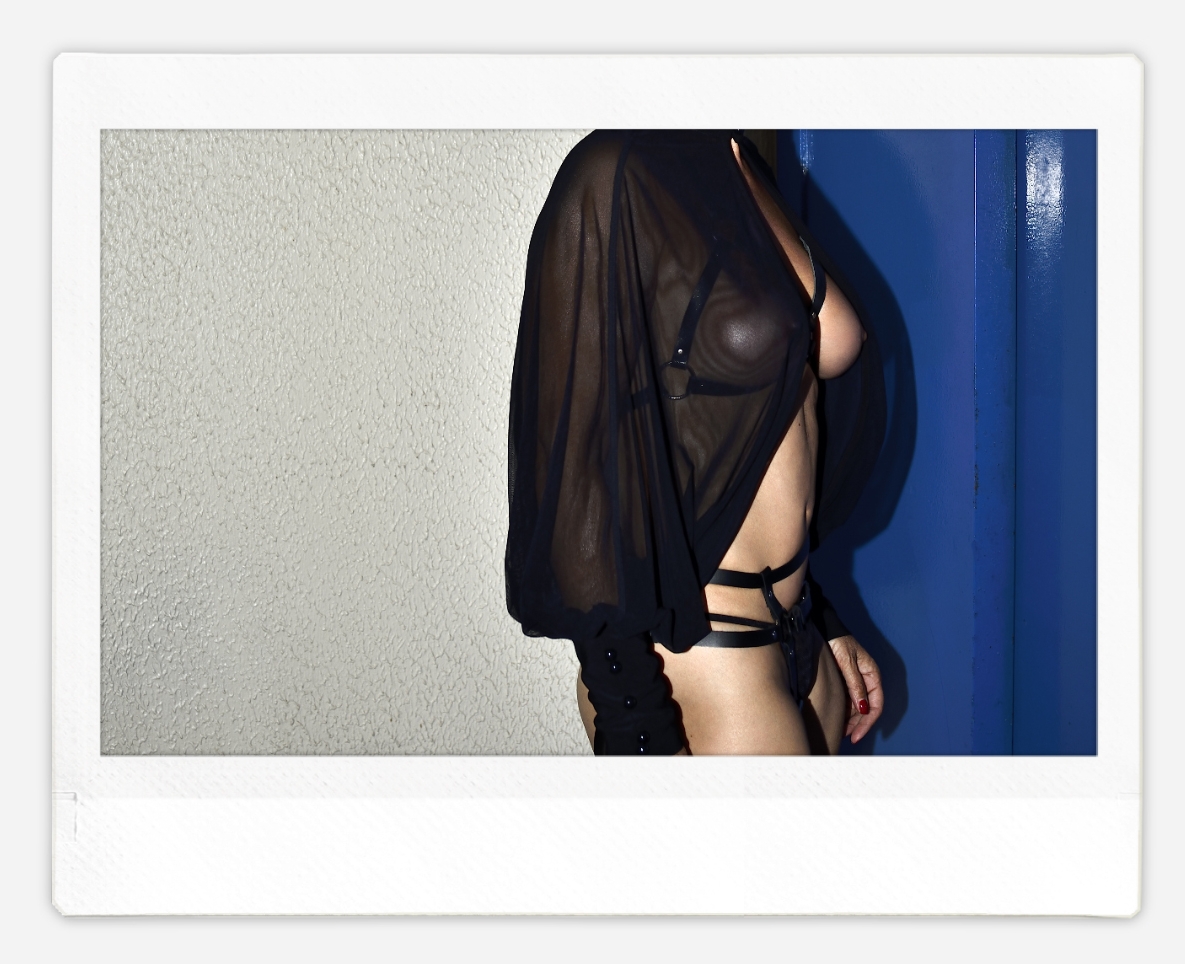 Maîtresse Lumina  portant un haut noir transparent et un bas noir à bretelles se tient près d'une porte bleue et d'un mur beige. Photo instantanée sur support Fujifim Instax Wide.