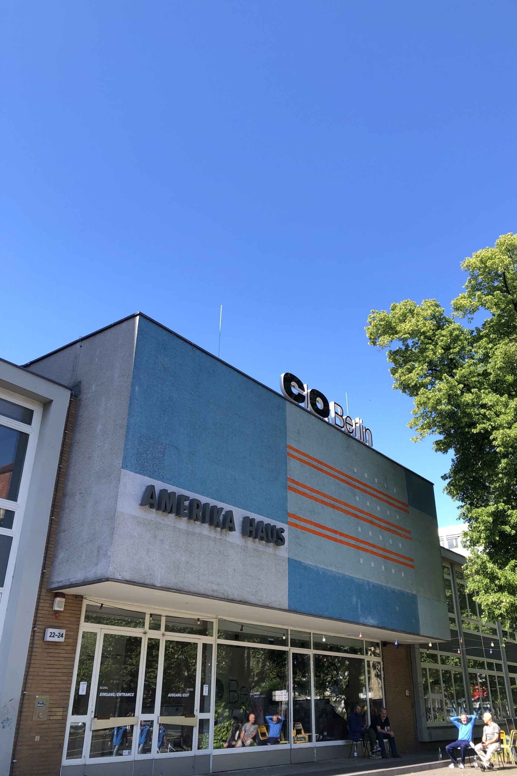 La photo montre l'Amerika Haus Berlin, un bâtiment moderne avec une façade bleue, des lignes horizontales orange et les mots « AMERIKA HAUS » et « C/O Berlin » sur le dessus, sur fond de ciel bleu clair et d'arbres verts.