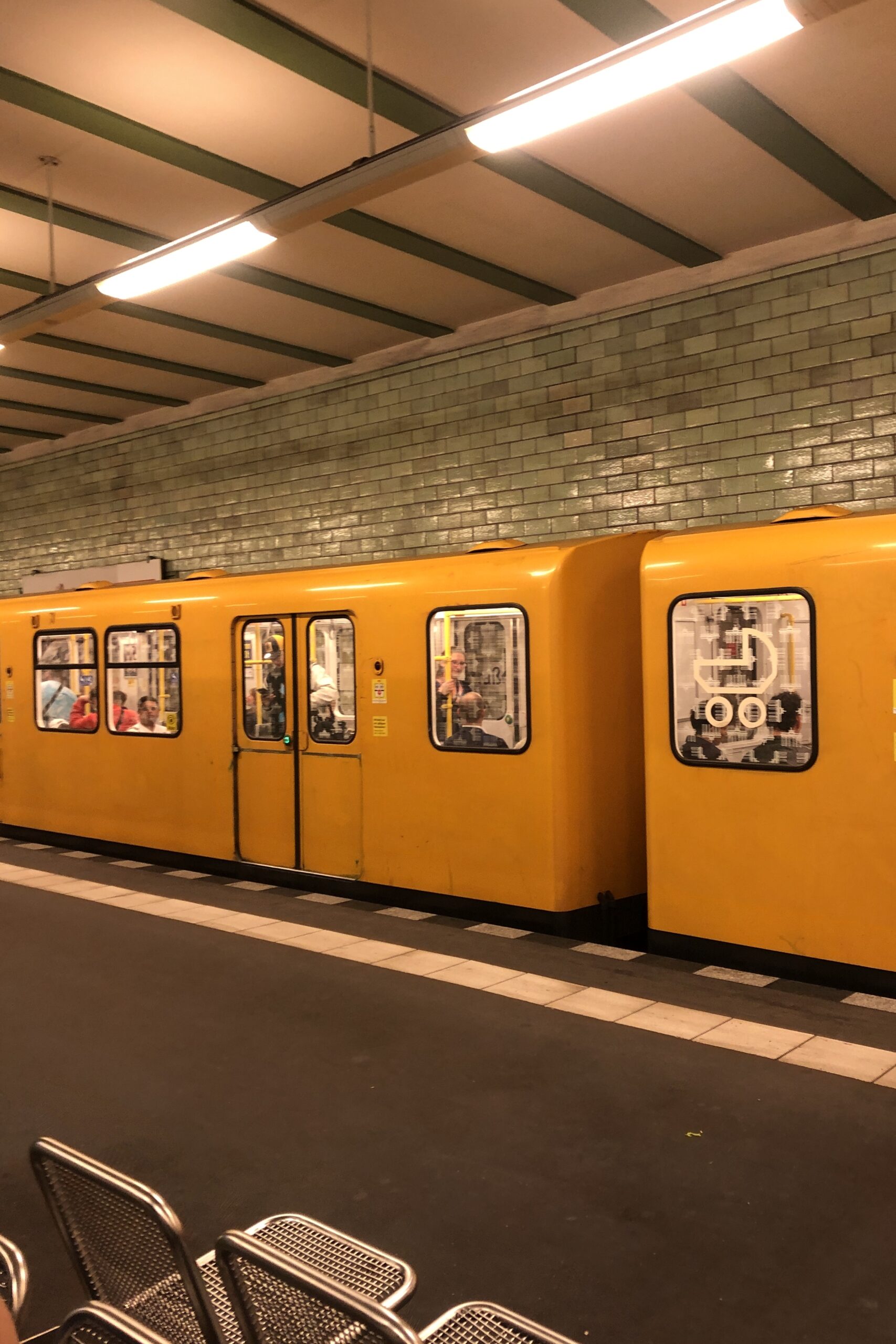 Une rame de métro jaune est arrêtée dans une station souterraine aux murs carrelés de vert. Plusieurs passagers sont visibles à l'intérieur, et des bancs métalliques vides apparaissent au premier plan.